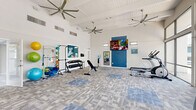 Located at 4581 South Kirkman Road in Orlando, Florida, this fitness center offers a spacious and thoughtfully designed workout environment across a single open floor. The gym's most striking architectural feature is its soaring vaulted ceiling, accented with exposed white wood paneling and outfitted with multiple large ceiling fans that ensure consistent airflow throughout the space. An entire wall of floor-to-ceiling windows and glass doors floods the interior with natural light while offering views of the surrounding outdoor area, creating an open and energizing atmosphere. The flooring consists of durable, patterned carpet tiles in neutral grey and blue tones that define different workout zones. The cardio section is well-appointed with elliptical machines, a treadmill, a stationary bike with an integrated display screen, and a recumbent bike. A dedicated strength training area features a full dumbbell rack, an adjustable weight bench, and a multi-function pull-up and dip station. Colorful stability balls, resistance bands, and yoga mats are neatly stored along one wall, supporting flexibility and functional fitness routines. A wall-mounted flat-screen television, a water station, and full-length mirrors complete the space, providing both entertainment and practical utility for a comprehensive workout experience.