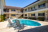 Located at 8722 Delgany Ave in the coastal Playa del Rey neighborhood of Los Angeles, this two-story multi-unit building is designed around a central courtyard that serves as the heart of the property. The layout forms a U-shaped configuration, with units on both floors oriented inward toward a pristine oval swimming pool surrounded by a generous poured-concrete deck. Lounge chairs and patio seating are positioned poolside, creating an inviting outdoor living area bathed in the abundant Southern California sunshine.

The building's exterior features a classic mid-century aesthetic, with a light yellow stucco facade, dark trim accents, and a low-pitched roof. Ground-floor units have direct access to the courtyard, while upper-floor units are served by open-air walkways with white metal railings, offering views over the pool area. Exterior stairways with concrete steps and white railings connect the two levels at multiple points throughout the courtyard.

Mature shrubs, small trees, and planted beds are integrated throughout the courtyard and along the building's base, lending greenery and a sense of privacy to the space. Large windows on each unit allow for natural light, while wall-mounted light fixtures provide illumination in the evenings. The overall ambiance is relaxed and resort-like, reflecting the laid-back coastal character of its Playa del Rey location.