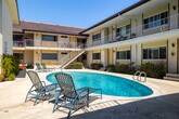 Located at 8722 Delgany Ave in the coastal Playa del Rey neighborhood of Los Angeles, this two-story multi-unit building is designed around a central courtyard that serves as the heart of the property. The layout forms a U-shaped configuration, with units on both floors oriented inward toward a pristine oval swimming pool surrounded by a generous poured-concrete deck. Lounge chairs and patio seating are positioned poolside, creating an inviting outdoor living area bathed in the abundant Southern California sunshine.

The building's exterior features a classic mid-century aesthetic, with a light yellow stucco facade, dark trim accents, and a low-pitched roof. Ground-floor units have direct access to the courtyard, while upper-floor units are served by open-air walkways with white metal railings, offering views over the pool area. Exterior stairways with concrete steps and white railings connect the two levels at multiple points throughout the courtyard.

Mature shrubs, small trees, and planted beds are integrated throughout the courtyard and along the building's base, lending greenery and a sense of privacy to the space. Large windows on each unit allow for natural light, while wall-mounted light fixtures provide illumination in the evenings. The overall ambiance is relaxed and resort-like, reflecting the laid-back coastal character of its Playa del Rey location.