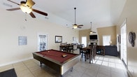 This single-floor space is designed with open-concept living in mind, seamlessly blending entertainment and relaxation into one expansive layout. The room's most striking architectural feature is its vaulted ceiling, which lends an airy, generous sense of height throughout the space. Multiple ceiling fans are suspended at varying levels, ensuring comfort across the entire area.

At the forefront of the room sits a full-size pool table with a rich burgundy felt surface, making it an immediate focal point and centerpiece for recreation. Behind it, a dining area accommodates a large table with multiple chairs, ideal for group gatherings. Further into the space, a bar-height table with bar stools provides an additional seating option.

The living area toward the rear of the room features a wall-mounted flat-screen television above a fireplace mantel, creating a cozy yet functional lounge zone. Large windows and a glass-paned exterior door flood the interior with natural light, reflecting off the neutral-toned tile flooring. The light-colored walls and open layout contribute to a bright, welcoming ambiance suited for both leisure and entertaining.