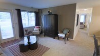 This single-floor space offers a thoughtfully designed living area that balances comfort and style. The living room serves as the focal point, featuring plush wall-to-wall carpeting that adds warmth underfoot and a soft, neutral foundation throughout. The room is anchored by a rich, dark-stained armoire that provides both storage and a striking visual centerpiece against a warm taupe accent wall. Natural light flows in generously through large windows dressed with dark curtain panels, offering a pleasant view of mature trees outside. The seating arrangement is carefully curated, combining a clean-lined upholstered accent chair with coordinating throw pillows and a woven rattan armchair with a cushioned seat, lending an earthy, layered texture to the space. A sleek, round glass-top coffee table rests on a striped area rug, adding a contemporary touch that contrasts nicely with the more traditional furnishings. Framed wall art and decorative accessories, including sculptural pieces atop the armoire, contribute to the polished, cohesive aesthetic. The overall ambiance is warm, inviting, and well-balanced, making this space well-suited for relaxed everyday living.