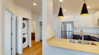 This single-floor residence showcases a clean, contemporary design with an open-concept layout that connects the living area to a well-appointed kitchen. The kitchen features a peninsula countertop with warm-toned surfaces, white upper cabinetry, dark lower cabinetry, and a full suite of stainless steel appliances, including a refrigerator, electric range, and microwave. A trio of striking black cone-shaped pendant lights hang above the peninsula, adding a modern aesthetic accent. Recessed lighting throughout ensures consistent, well-distributed illumination.

Light hardwood-style flooring flows through the main living and kitchen areas, while the bedroom features plush wall-to-wall carpeting. The bedroom benefits from natural light through a window fitted with horizontal blinds, providing exterior views. A dedicated in-unit laundry area houses a stacked washer and dryer, offering everyday convenience. The bathroom presents a clean, functional design with a grey vanity, neutral stone-toned countertop, an undermount sink with brushed nickel fixtures, and a large mirror above. A walk-in closet provides additional storage. The overall ambiance is bright and modern, emphasizing neutral tones and practical finishes throughout.