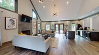 Located at 16001 Summerwood Ln in Alpharetta, Georgia, this single-floor space delivers an impressive blend of architectural character and modern comfort. The centerpiece of the main living area is a dramatic vaulted ceiling with distinctive arched windows that draw in abundant natural light and frame views of surrounding greenery. Dark wood crown molding and exposed beams accent the soaring roofline, lending warmth and definition to the airy interior.

Rich hardwood flooring flows throughout the space, anchoring the open-concept layout. A floor-to-ceiling tile fireplace serves as a commanding focal point along one wall, complemented by a wall-mounted flat-screen television above. Multiple seating arrangements—including neutral-toned sofas, accent chairs, and a substantial rustic wood coffee table—create a welcoming and versatile environment.

A sputnik-style chandelier adds a contemporary touch overhead, while track lighting provides additional ambient illumination. Adjacent to the main lounge is a functional bar and refreshment area featuring a raised countertop with leather barstools, dark cabinetry, a beverage refrigerator, and a coffee station set against textured wallcovering.

French doors open directly to an outdoor space, enhancing the connection between interior and exterior. The overall ambiance is sophisticated yet approachable, making this a well-appointed communal or event space.