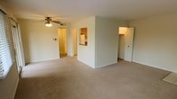 This single-floor residence offers a straightforward and functional layout designed for comfortable everyday living. The main living area is finished with wall-to-wall carpet in a neutral tone, creating a warm and inviting atmosphere. A ceiling fan with integrated lighting is centrally positioned, providing both air circulation and ambient illumination throughout the space. The living area connects seamlessly to the rest of the unit, with a pass-through window opening into the kitchen, which features wooden cabinetry and a white refrigerator, allowing for easy interaction between the two spaces. Adjacent to the kitchen pass-through is a doorway leading further into the unit, with light-colored flooring visible beyond the threshold. On the opposite side of the living area, a generously sized closet with a built-in hanging rod offers ample storage. The walls throughout are painted in a clean, light neutral tone, contributing to a bright and open feel. The overall design prioritizes practicality and ease of use, making this a well-organized space suited for a variety of living arrangements.