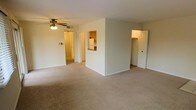 This single-floor residence offers a straightforward and functional layout designed for comfortable everyday living. The main living area is finished with wall-to-wall carpet in a neutral tone, creating a warm and inviting atmosphere. A ceiling fan with integrated lighting is centrally positioned, providing both air circulation and ambient illumination throughout the space. The living area connects seamlessly to the rest of the unit, with a pass-through window opening into the kitchen, which features wooden cabinetry and a white refrigerator, allowing for easy interaction between the two spaces. Adjacent to the kitchen pass-through is a doorway leading further into the unit, with light-colored flooring visible beyond the threshold. On the opposite side of the living area, a generously sized closet with a built-in hanging rod offers ample storage. The walls throughout are painted in a clean, light neutral tone, contributing to a bright and open feel. The overall design prioritizes practicality and ease of use, making this a well-organized space suited for a variety of living arrangements.