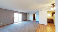 This single-floor space offers a clean, functional layout designed for comfortable everyday living. The main living area is generously sized, featuring plush wall-to-wall carpeting that transitions seamlessly into warm hardwood flooring, creating a clear visual distinction between living and adjoining spaces. A textured ceiling adds subtle depth overhead, while neutral-toned walls in soft grey and white provide a versatile backdrop throughout.

A sliding glass door dominates one wall of the living area, flooding the space with natural light and offering direct access to an outdoor balcony — a standout feature that enhances both the brightness and the sense of openness within the room. The balcony view hints at an elevated position, making it a desirable spot for outdoor enjoyment.

HVAC vents are integrated into the walls, ensuring efficient climate control throughout. A doorway visible from the main living area leads further into the space, suggesting additional rooms beyond, including what appears to be a closet with louvered doors. The overall ambiance is bright, open, and move-in ready, with a straightforward layout that lends itself well to a variety of furniture arrangements and personal styling preferences.