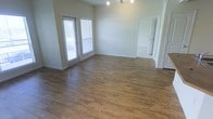This single-story space presents a bright, open main living area highlighted by warm-toned hardwood-style flooring that extends throughout. The room is finished with crisp white trim and neutral-colored walls, creating a clean and versatile backdrop. Natural light filters in through two windows dressed with horizontal blinds, including what appears to be a glass-paneled door providing access to an outdoor area. A multi-bulb ceiling light fixture anchors the space above, offering ample overhead illumination. The layout feels open and unobstructed, allowing for flexible furniture arrangements. White baseboards frame the perimeter neatly, adding a polished finishing detail. A doorway visible in the corner leads to an adjoining room, suggesting a connected and functional floor plan. The transition from the hardwood-style flooring to what appears to be carpet in the adjacent room indicates distinct zones within the unit. Overall, the space presents a straightforward, well-maintained interior with a neutral aesthetic that accommodates a variety of uses and personal styles.