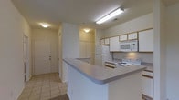 This single-story unit is centered around a practical, open-concept kitchen that flows seamlessly into the entryway and adjacent living areas. The kitchen is equipped with white appliances, including a refrigerator, electric range, and over-the-range microwave, complemented by light wood-trimmed upper cabinetry providing ample storage. A peninsula counter with a gray laminate surface extends into the space, serving as a natural divider between the kitchen and the surrounding areas while offering additional workspace. Overhead fluorescent lighting illuminates the kitchen efficiently, while flush-mount ceiling fixtures carry light into the connecting spaces. The flooring transitions between light ceramic tile in the kitchen and entryway areas, adding a clean, neutral foundation throughout. The entryway, visible from the kitchen, features a solid paneled front door and straightforward access into the unit. The overall design is functional and straightforward, with white walls and neutral finishes creating a bright, cohesive aesthetic throughout the space.