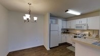 This single-story space showcases a functional and cohesive open layout connecting the kitchen and dining area. The kitchen is outfitted with white cabinetry that extends to the upper walls, providing ample storage space. A light-colored countertop runs along the base cabinets, complemented by white appliances including a full-size refrigerator, a range, and a built-in microwave. Recessed fluorescent lighting above the kitchen workspace ensures a well-lit cooking environment. A patterned area rug adds a subtle decorative touch near the kitchen entrance.

The adjacent dining area flows naturally from the kitchen and features dark hardwood-style flooring that extends throughout the visible space, grounding the room with warmth. A three-bulb brushed nickel chandelier serves as a focal point overhead, lending a classic and inviting atmosphere to the dining space. The walls are finished in a neutral tone, and the textured ceiling adds a traditional residential character. Clean white baseboards provide a crisp contrast to the darker flooring, contributing to the overall tidy and well-maintained feel of the space.