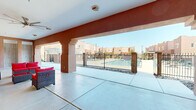 This inviting outdoor amenity space showcases a distinctly Southwestern architectural style, characterized by warm terracotta and adobe-toned stucco exteriors, decorative vigas, and earthy tones that echo the surrounding desert landscape. At the heart of the space is a generously sized swimming pool with clear, blue water, surrounded by a wide concrete pool deck furnished with chaise lounges for sunbathing. The pool area is framed by an ornamental wrought-iron fence with decorative stone-filled planters at its base, providing both security and aesthetic appeal. Two covered gazebo structures flank the pool deck, offering shaded retreats ideal for relaxing or gathering. A spacious covered patio anchors one side of the courtyard, featuring a vaulted ceiling with a large ceiling fan, polished concrete flooring, and comfortable wicker lounge seating with bold red cushions. This sheltered area also includes outdoor shower stalls and direct access to the building interior. The overall ambiance is bright and open, bathed in abundant natural sunlight beneath clear skies. The entire courtyard is enclosed within a secure perimeter fence, creating a private, resort-like retreat with a cohesive Southwestern design aesthetic throughout.