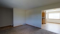 Located at 5368 Big Tyler Rd in Cross Lanes, West Virginia, this two-floor property presents a straightforward and functional layout designed for comfortable everyday living. The main living area features wall-to-wall carpeting in a neutral tone, creating a warm and understated foundation that adapts easily to a variety of furnishing styles. Light-colored walls throughout contribute to a clean, open atmosphere, while a floor-level ventilation register indicates a forced-air heating and cooling system for year-round climate control. A thermostat is visibly mounted on the wall, offering convenient access to temperature management. The living space flows directly into the kitchen, which is visible through a connecting doorway. The kitchen features warm honey-toned wood cabinetry, dark countertops, and tile-style flooring that contrasts with the carpeted living area, providing a practical and easy-to-maintain surface. The open sightline between the two spaces supports a connected, cohesive feel. Spanning two floors, the property offers additional space above, suitable for bedrooms or other private living areas. The overall ambiance is clean, neutral, and move-in ready.
