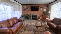 This single-floor space exudes a warm, classic charm anchored by a striking brick fireplace that serves as the focal point of the living area. The fireplace is framed by rich brick detailing and topped with a large flat-screen television, making it both a functional and decorative centerpiece. Earthy terracotta-toned walls lend a cozy, inviting ambiance throughout the room, complemented by crown molding that adds a touch of refined character. The living area is furnished with generous leather sofas and a matching armchair in a rich caramel tone, arranged around an ornate wooden coffee table with a glass top. A detailed area rug anchors the seating arrangement, adding texture and warmth underfoot. Natural light filters softly into the room through a window dressed with delicate floral curtains, brightening the space without overpowering its cozy feel. Lush indoor plants are thoughtfully placed throughout, introducing a refreshing natural element. The walls are tastefully adorned with a curated arrangement of framed artwork, adding personality and visual interest. Overall, the space presents a well-appointed, comfortable environment with a traditional aesthetic and attention to detail.