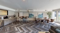 Located at 10343 San Diego Mission Rd in San Diego, CA, this thoughtfully designed single-floor community clubhouse offers a stylish and functional gathering space for residents. The interior is anchored by a sophisticated lounge area featuring a sleek gas fireplace set against a floor-to-ceiling tile surround, with a wall-mounted flat-screen TV above. Vibrant teal mid-century modern accent chairs and a plush gray sofa create an inviting seating arrangement, while patterned herringbone carpet tiles add texture and visual interest throughout. Large floor-to-ceiling windows flood the lounge with generous natural light and offer views of the surrounding landscaped grounds. A dedicated meeting and co-working area provides high-top and standard seating arrangements, ideal for both collaborative and individual use, complemented by contemporary wall art and a mini-fridge for added convenience. The entry zone is defined by striking black-framed glass partition walls and doors, lending an industrial-chic aesthetic that connects the spaces with transparency and openness. Lush potted tropical plants enhance the welcoming atmosphere. Recessed lighting throughout ensures a clean, modern finish, while the overall neutral palette of whites, grays, and warm wood tones creates a cohesive and polished ambiance suited for both social and productive use.