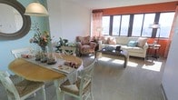 This inviting living space showcases an open-concept design that seamlessly blends dining and lounging areas. Large windows flood the room with natural light, creating a bright and airy atmosphere. The space is furnished with an eclectic mix of pieces, including a round dining table with white chairs, a floral-patterned armchair, and a comfortable sofa. A striking orange accent wall adds warmth and vibrancy to the neutral backdrop. The room's decor incorporates various textures, from woven chair seats to plush cushions, enhancing its cozy appeal. A glass coffee table and strategically placed lamps contribute to the room's functional yet stylish ambiance. The carpeted floor adds comfort underfoot, while carefully chosen accessories, such as potted plants and decorative items, personalize the space. This versatile area appears ideal for both casual dining and relaxed socializing, embodying a harmonious blend of comfort and contemporary design.