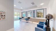 Located at 1231 Arya Way in Ocoee, Florida, this contemporary beauty salon offers a clean, inviting environment designed with both functionality and aesthetics in mind. The single-floor layout makes efficient use of its compact footprint, accommodating multiple service zones within an open, flowing floor plan.

The main styling area is a standout feature, showcasing a row of illuminated vanity mirrors framed against a blue tile accent wall. Warm wood-toned cabinetry runs beneath the mirrors, housing integrated wash basins and ample storage. Blue upholstered styling chairs complement the cool, calming color palette throughout the space.

A dedicated nail care station occupies a quieter corner, complete with a manicure table, client seating, and a pedicure chair. A wall-mounted nail polish rack displays an organized selection of color options, adding a decorative and practical touch to the area.

The salon also features a hair drying station with two plush blue chairs positioned beneath hood dryers, providing a comfortable setting for clients. Elegant framed floral artwork adorns nearby walls, contributing to the spa-like ambiance. Large windows along one wall invite abundant natural light, offering views of the surrounding greenery. Light-toned tile flooring and white walls throughout the space reinforce the fresh, modern aesthetic that defines this well-designed salon.