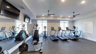 This modern fitness center is thoughtfully designed to support a well-rounded workout routine. The space is anchored by a lineup of cardio equipment, including multiple treadmills, ellipticals, a stair climber, and a stationary bike, with several machines featuring individual digital display screens for tracking performance. Large floor-to-ceiling windows span the far wall, flooding the interior with natural light and offering views to the outdoors, creating an open and energizing atmosphere. Recessed lighting and ceiling fans are positioned throughout, ensuring the space remains well-lit and comfortably ventilated during workouts. Wall-mounted flat-screen televisions provide entertainment options for users during their sessions. The flooring consists of durable, cushioned gym-grade material suited for high-traffic fitness use. The neutral color palette of soft grays and whites contributes to a clean, focused environment. The layout is organized and spacious, allowing for comfortable movement between equipment. Overall, the fitness center delivers a practical and inviting setting for cardiovascular training and general fitness activity.