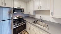 This single-floor unit offers a clean, modern layout designed for comfortable everyday living. The open living and dining area is finished with light-toned hardwood-style flooring and neutral walls, creating a bright and airy atmosphere. A ceiling fan overhead provides added comfort, while a large window with vertical blinds allows natural light to fill the space. A built-in countertop peninsula connects the living area to the kitchen, offering a convenient transition between the two spaces.

The kitchen is thoughtfully designed with white shaker-style cabinetry extending to the ceiling, providing ample storage. Grey laminate countertops complement the stainless steel appliances, which include a gas range, over-the-range microwave, refrigerator, and a single-basin sink. The clean lines and neutral palette give the kitchen a functional yet polished look.

A dedicated hallway leads to the bedroom and bathroom. The bedroom features plush carpeting, soft neutral walls, and a window with vertical blinds that invites natural light. A closet off the hallway offers convenient storage. The bathroom is finished with a white vanity, grey countertop, a large mirror, and wood-style flooring, maintaining the cohesive aesthetic found throughout the unit. Overall, the space presents a fresh, move-in-ready environment with consistent modern finishes.