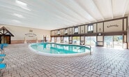 Located at 3710 Inverrary Drive in Lauderhill, Florida, International Village — "The Jewel of Inverrary" — is a distinctive recreational facility defined by its charming Tudor-inspired architecture. The indoor pool area is a standout feature, showcasing a generously sized swimming pool with crystal-clear turquoise water, set beneath soaring ceilings with exposed white beam detailing. The enclosure is framed by dark wood timber accents, brick wainscoting, and a row of large windows along the upper mezzanine level that flood the space with natural light and offer views of the surrounding outdoor area. Paver brick flooring runs throughout, lending a warm, resort-like ambiance. Blue lounge chairs are thoughtfully arranged poolside for relaxation. A dedicated spa area is also found within the enclosure, featuring its own hot tub, wall sconce lighting, and decorative ironwork. Classical statuary adds an elegant, artistic touch near the entrance, which bears the community's signature branding. Large sliding glass doors seamlessly connect the indoor facility to an expansive outdoor pool deck, where additional lounge seating, lush landscaping, and open-sky views create a vibrant aquatic retreat for residents and guests alike.