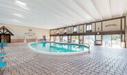 Located at 3710 Inverrary Drive in Lauderhill, Florida, International Village — "The Jewel of Inverrary" — is a distinctive recreational facility defined by its charming Tudor-inspired architecture. The indoor pool area is a standout feature, showcasing a generously sized swimming pool with crystal-clear turquoise water, set beneath soaring ceilings with exposed white beam detailing. The enclosure is framed by dark wood timber accents, brick wainscoting, and a row of large windows along the upper mezzanine level that flood the space with natural light and offer views of the surrounding outdoor area. Paver brick flooring runs throughout, lending a warm, resort-like ambiance. Blue lounge chairs are thoughtfully arranged poolside for relaxation. A dedicated spa area is also found within the enclosure, featuring its own hot tub, wall sconce lighting, and decorative ironwork. Classical statuary adds an elegant, artistic touch near the entrance, which bears the community's signature branding. Large sliding glass doors seamlessly connect the indoor facility to an expansive outdoor pool deck, where additional lounge seating, lush landscaping, and open-sky views create a vibrant aquatic retreat for residents and guests alike.