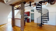 Located at 1329 Jefferson St in Lynchburg, Virginia, this remarkable two-story loft is a beautifully converted industrial space that masterfully blends historic character with modern comfort. Original heavy timber posts and beams dominate the interior, serving as striking architectural reminders of the building's industrial past, while preserved mill machinery — including large cast-iron wheels mounted overhead — add a bold, authentic industrial aesthetic throughout.

Warm hardwood floors run continuously across both levels, complementing the crisp white walls and abundant natural light that filters through large windows. The open main level centers around a spacious living area framed by the signature timber columns, with a ceiling fan overhead and easy access to a well-appointed galley kitchen. The kitchen features rich cherry cabinetry, granite countertops, stainless steel appliances, and hardwood flooring. A clean, tiled full bathroom with a tub/shower combo and cherry vanity provides functional comfort.

A sleek black metal spiral staircase leads to the upper level, where exposed timber beams follow the dramatic roofline, creating an intimate yet airy atmosphere. A bedroom on this level features white-painted exposed brick walls and a generous closet. Throughout the space, the combination of reclaimed materials, soaring ceilings, and industrial heritage creates a distinctive and memorable living environment.