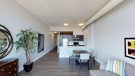 This single-floor space showcases a thoughtfully designed open-concept layout that seamlessly integrates the kitchen, dining, and living areas into one cohesive environment. The kitchen is outfitted with dark espresso cabinetry, stainless steel appliances including a refrigerator, microwave, and range, along with a bright white peninsula that provides additional prep space and serves as a natural divider between the kitchen and dining areas. Pendant lighting above the peninsula adds a modern, refined touch. The dining area comfortably accommodates a rectangular table with upholstered chairs, positioned to take full advantage of the open flow. Rich, dark hardwood-style flooring runs throughout the space, grounding the interior with warmth and texture, while the crisp white walls and ceilings create a bright, airy atmosphere. Architectural elements such as a ceiling soffit with integrated ductwork lend an industrial character to the otherwise clean, contemporary design. The living area, partially visible, features a wall-mounted flat-screen television and a large potted plant that introduces a touch of greenery. Recessed and flush-mount ceiling lighting ensures the space is well-lit throughout, enhancing the overall modern and functional ambiance.