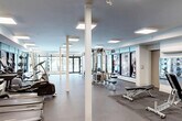 Located at 11025 26 Ave NW in Edmonton, Alberta, this well-appointed fitness facility offers a generous, single-floor exercise space designed to support a comprehensive range of workout routines. The exercise room is thoughtfully laid out, with dedicated zones for cardiovascular training, free weights, and strength equipment. Cardio machines — including treadmills and ellipticals — are positioned along one side of the room, while a full dumbbell rack and adjustable benches occupy the free weights area. Multi-station cable machines and a power rack further expand the strength training options available. The space features durable rubber flooring throughout, providing both safety and comfort underfoot. Large floor-to-ceiling windows along the perimeter flood the room with abundant natural light and offer views of the surrounding building exterior. Recessed panel lighting supplements the natural light, ensuring the space remains bright and well-illuminated. White structural columns are integrated into the open layout without interrupting the flow of the space. Wall-mounted mirrors enhance the sense of openness. A storage unit and a mini-split climate control system are also present, contributing to the overall comfort and functionality of this well-equipped fitness environment.