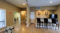 This single-floor space offers a cohesive, open layout designed for comfortable everyday living. The interior is finished with warm taupe and neutral-toned walls throughout, complemented by light hardwood-style flooring that runs seamlessly across the main areas, creating a sense of continuity and spaciousness.

A standout feature is the breakfast bar, which includes a light-colored countertop and is paired with two dark upholstered bar stools, making it an ideal spot for casual dining or conversation. Recessed lighting is installed throughout the ceiling, providing consistent and well-distributed illumination across the space.

The open hallway connects to multiple rooms, including a dining area visible through an elegantly arched doorway, where a dark wood table and chairs can be seen. This architectural detail adds a touch of character to the otherwise clean and modern interior. A second doorway leads to an additional room, offering flexible use of space.

White trim and baseboards provide a crisp contrast against the warmer wall tones, while framed wall art adds decorative touches throughout. HVAC vents are strategically placed in both the ceiling and walls, ensuring efficient climate control. Overall, the space presents a tidy, functional, and welcoming environment with a consistent and polished aesthetic.