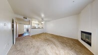 This two-floor residence in Pleasanton, California is designed with a practical, open layout that maximizes usable space throughout. The main living area is generously sized, finished with wall-to-wall neutral carpeting and bright white walls that lend a clean, airy feel. A ceiling fan anchors the room, and a sliding glass door provides access to an outdoor area, allowing natural light to flow in. The kitchen is positioned adjacent to the living space, separated by a raised peninsula counter that creates a natural divide while maintaining an open connection between the two areas. White cabinetry reaches toward the ceiling, complemented by wood-look flooring, a stainless steel refrigerator, and a full suite of appliances including a range with overhead ventilation. A staircase leads to the upper floor, where carpeted bedrooms offer quiet, comfortable retreats. One bedroom features multiple windows overlooking a green outdoor setting, filling the room with natural light. The bathroom is well-appointed with a vanity featuring a stone-tone countertop, a tub-shower combination, and accessibility grab bars throughout. Wood-look flooring carries through the bathroom, providing a cohesive finish across the home's hard-surface areas.