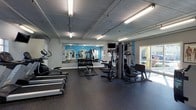 This single-level fitness center offers a comprehensive workout environment designed to accommodate a range of exercise routines. The space is outfitted with a row of treadmills along one wall, providing dedicated space for cardiovascular training. A multi-station cable machine and additional strength equipment occupy the central and far areas of the room, catering to resistance and functional training needs. A dumbbell rack with a flat bench sits prominently in the middle of the space, offering a full range of free weights for various exercises. The flooring consists of durable dark rubber matting, which provides both safety and noise reduction throughout. Bright blue accent walls add a vibrant, energizing atmosphere, while large full-wall mirrors on one side enhance the sense of depth and allow users to monitor their form. Overhead fluorescent lighting ensures the space is well-lit, and a wall-mounted television offers entertainment during workouts. A large window along one wall introduces natural light and views of the outdoors, contributing to an open and inviting environment. The overall layout is practical and efficiently organized.