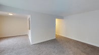 This single-floor space offers a clean, straightforward layout designed for practical, comfortable living. The interior is characterized by neutral, light gray walls and smooth white ceilings that create a bright and airy atmosphere throughout. Plush wall-to-wall carpeting in a warm beige tone runs continuously across the main living areas, adding a sense of softness and comfort underfoot. The open floor plan allows for a natural flow between spaces, with clearly defined areas separated by partial walls and open doorways that maintain a sense of connectivity without sacrificing structure. White baseboards provide a crisp contrast against the carpeting, contributing to the overall clean and well-maintained aesthetic. The layout includes multiple interconnected rooms, with passageways that lead to additional areas of the space, suggesting a thoughtful and functional arrangement. Recessed wall outlets and light switches are positioned throughout, reflecting practical design considerations. The overall ambiance is calm and understated, with a neutral color palette that provides a versatile backdrop suitable for a variety of interior styles and furnishings. The space presents a move-in ready environment with a modest, tidy character.