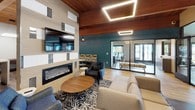 This single-floor communal space is thoughtfully designed to blend comfort and functionality in an open, contemporary layout. Warm wood-plank ceilings with exposed beams run throughout, complemented by light hardwood flooring and bold teal accent walls that add a vibrant pop of color. Geometric LED light fixtures are recessed into the ceiling, providing modern, ambient illumination alongside track lighting over the kitchen area.

The kitchen is well-equipped with a long central island featuring white quartz countertops, gray cabinetry, stainless steel appliances including a refrigerator and microwave, and a subway tile backsplash. A pendant light anchors the island as a focal point.

The lounge area is anchored by a sleek linear electric fireplace set into a textured feature wall, topped by a large wall-mounted television. Comfortable seating including a sectional sofa, armchairs, and a patterned area rug creates an inviting gathering space. A dining table with modern chairs provides additional seating nearby.

Floor-to-ceiling glass sliding doors open to views of an outdoor pool, flooding the space with natural light and seamlessly connecting the interior to the outdoor amenity area.