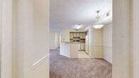 This single-story unit offers a practical and comfortable layout across three bedrooms and two full bathrooms. The open-concept living and kitchen area forms the heart of the home, where light-toned walls and warm carpet flooring create a welcoming atmosphere. The kitchen features a peninsula with a countertop overhang, light wood cabinetry, tile backsplash, tile flooring, and white appliances including an electric range and refrigerator. A pendant light fixture above the peninsula adds a subtle design touch.

The living area flows seamlessly from the kitchen and features a sliding glass door that opens to a private balcony, providing natural light and outdoor views of the surrounding greenery and neighboring buildings. A ceiling fan with a light fixture overhead adds comfort to the space.

Each of the three bedrooms is carpeted and finished with the same warm, neutral tones. Two bedrooms feature ceiling fans and windows with slatted blinds that bring in natural light and views of lush trees outside. The primary bedroom is a comfortable size and shares the same design consistency.

Both bathrooms are finished with light tile flooring and include full tub-shower combinations with white tile surrounds and chrome fixtures, providing clean and functional spaces. A hallway connects all rooms, completing the efficient and cohesive floor plan.