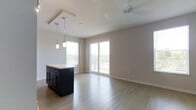 This single-floor space offers an open-concept layout that seamlessly connects the kitchen and living areas, creating a bright and airy atmosphere. The kitchen features a dark-cabinetry island topped with a light stone countertop, accented by a trio of modern pendant lights that hang at varying heights, adding a contemporary touch. Large windows allow abundant natural light to pour in, enhancing the warmth of the light wood-style flooring that extends throughout the space. The living area is anchored by a ceiling fan, providing both comfort and functionality. A full-height sliding glass door leads to an outdoor balcony, offering views of the surrounding area and creating a smooth indoor-outdoor connection. The neutral wall tones complement the clean lines and modern finishes found throughout, lending the space a versatile and inviting ambiance. The open layout and well-placed lighting make this a practical and stylish space well-suited for everyday living and entertaining alike.