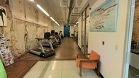 This single-level fitness space offers a functional and eclectic workout environment that blends industrial aesthetics with practical design. The room is characterized by its exposed ceiling ductwork and overhead fluorescent strip lighting, which runs the length of the space and provides consistent, even illumination throughout. One wall retains a distinctly raw, weathered texture, contributing to the space's urban industrial character, while the opposite side features clean white walls and modern glass-paneled partitions that allow for visual transparency between areas.

The flooring transitions from dark hardwood in the main exercise area to light tile near the entrance, subtly delineating zones within the space. A row of cardio equipment, including treadmills and elliptical machines, lines one side of the room, while additional strength and cable equipment is positioned toward the far end near a teal-painted door that serves as a focal point at the rear. A wooden-framed orange chair is positioned along the right wall, offering a seating area for rest between workouts. A colorful piece of wall art adds a vibrant, creative touch to the otherwise utilitarian environment, enhancing the overall ambiance of the space.