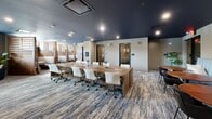 Located at 51 S Broadway in White Plains, NY, this professionally designed coworking space offers a well-appointed, single-floor workspace with a diverse range of functional areas to accommodate various working styles. The interior is anchored by a striking deep navy ceiling that runs throughout, lending a polished, contemporary atmosphere complemented by recessed lighting and textured, multi-toned carpeting.

The centerpiece of the space is a large, solid walnut communal work table lined with white ergonomic chairs, providing ample seating for collaborative work or focused individual tasks. Adjacent to this main area, a lounge zone features a curved navy sofa, accent chairs, and a round wooden coffee table — ideal for informal meetings or relaxed work sessions. Lush indoor plants add a refreshing, organic touch throughout.

A row of semi-private booth seating with slatted wood dividers offers quieter, more secluded work areas, while a dedicated lounge nook with a tufted banquette, round tables, and an eclectic wall clock display creates a welcoming social corner. Open shelving with warm wood tones frames the reception-style entry area, and several private rooms with glass-paneled doors provide enclosed spaces for calls or focused work. Natural light filters in through multiple windows, enhancing the overall sense of openness and professionalism throughout this thoughtfully curated workspace.