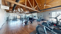 Located at 18303 Cottonwood Drive in Parker, Colorado, this single-level fitness facility offers a well-appointed and expansive workout environment defined by striking architectural character. The space is anchored by soaring vaulted ceilings with exposed wooden tongue-and-groove paneling and substantial timber trusses with visible steel gusset plate connections, lending the interior a warm, lodge-inspired aesthetic. Hardwood flooring runs throughout, complementing the natural wood tones overhead.

The facility is organized into distinct functional zones. The main cardio area features a row of treadmills, ellipticals, stair climbers, and stationary bikes, positioned near large windows that admit generous natural light and views of the surrounding landscape. Rowing machines occupy an open stretch of floor, providing additional cardio options.

The strength training area includes cable machines, free weights, adjustable benches, and a dumbbell rack. A dedicated studio space features floor-to-ceiling mirrors, a ballet barre along one wall, TRX suspension equipment, resistance bands, and medicine balls, suitable for group fitness or individual training sessions. A wall-mounted flat-screen TV and a small beverage refrigerator add functional amenities throughout. Large ceiling fans and exposed ductwork contribute to the facility's open, athletic ambiance.