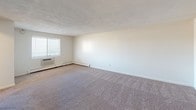 Located at 405 Quaker Ln in West Warwick, Rhode Island, this single-floor unit offers a practical and comfortable layout designed for everyday living. The spacious living area is finished with neutral-toned wall-to-wall carpeting, textured ceilings, and a ceiling fan with integrated light, while a window with blinds allows natural light to filter in alongside a baseboard heating system and wall-mounted air conditioning unit. An adjacent room, also carpeted, provides additional flexible living or dining space with its own window and ceiling fan. The kitchen is efficiently arranged with white cabinetry featuring black hardware, light stone-patterned countertops, a double stainless steel sink, and durable wood-look flooring, along with ample upper and lower cabinet storage. The bedroom is generously sized with plush carpeting, a built-in closet with sliding doors offering organized storage, and direct access to the bathroom. The full bathroom is appointed with wood-look vinyl flooring, a combined tub and shower unit, a toilet, a vanity with a white countertop, and a baseboard heater. Throughout the unit, the clean, neutral color palette and functional finishes create a bright and move-in ready environment.