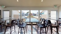This contemporary clubhouse is thoughtfully designed to offer a welcoming and functional community gathering space. The main lounge area immediately impresses with its soaring vaulted ceilings supported by exposed beams, large pendant light fixtures, and an abundance of natural light that flows through generous windows. Comfortable seating arrangements — including a plush gray sectional and leather accent chairs — are anchored by a textured area rug, creating an inviting atmosphere for relaxation or socializing. A striking floor-to-ceiling fireplace with a white shiplap surround serves as a central focal point, complemented by a wall-mounted television above.

The flooring transitions seamlessly between large-format dark tile and a richly varied reclaimed wood plank surface, adding warmth and visual interest throughout. The fully equipped kitchen features dark espresso cabinetry extending to the ceiling, light quartz countertops, a subway tile backsplash, and a suite of stainless steel appliances including a gas range, refrigerator, and dishwasher — ideal for hosting events or catering needs.

A dedicated dining area with a high-top communal table and bar-height seating is positioned adjacent to a wall of large windows, offering direct views of the outdoor pool and surrounding amenity spaces, filling the room with natural light and a vibrant outdoor connection.
