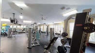 This fitness center offers a spacious, single-floor layout designed to accommodate a wide range of workout needs. The facility is divided into two distinct zones: a strength training area and a dedicated cardio section, each thoughtfully arranged to maximize usability and flow.

The strength training area is generously stocked with a variety of cable machines, selectorized weight stations, free-weight benches, and plate-loaded equipment from brands such as Paramount. Full-length wall mirrors run throughout, enhancing the sense of openness while allowing users to monitor their form. Multiple windows along the perimeter walls invite natural light and provide views of the surrounding greenery, creating an energizing atmosphere.

The cardio zone features treadmills, stair climbers, stationary bikes, and additional circuit machines arranged along mirrored walls. Ceiling-mounted flat-screen televisions are positioned throughout for entertainment during workouts. A ceiling fan aids in air circulation, contributing to overall comfort.

Underfoot, durable rubber flooring covers the entire space, providing safety and sound absorption. Recessed and flush-mounted fluorescent lighting supplements the natural light, ensuring the facility is well-lit at all times. The warm yellow-toned walls add a welcoming, clean aesthetic throughout.