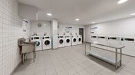 Located at 2570 Kingston Rd in Toronto's Kingston Road corridor, this shared laundry facility offers a clean, well-organized space designed for practical, high-volume use. The room is finished with white square wall tiles from floor to ceiling, contributing to a bright and hygienic environment, while large-format light grey floor tiles provide a durable and easy-to-maintain surface throughout.

The facility is equipped with a row of front-loading commercial washing machines and a matching set of commercial dryers, each unit featuring individual digital controls and card-based payment systems for convenient operation. A stainless steel utility sink is mounted along one wall, providing a dedicated handwashing or pre-treatment station. Generous stainless steel folding tables are positioned centrally and along the perimeter, offering ample workspace for sorting and folding laundry.

Recessed ceiling lights illuminate the space evenly, ensuring clear visibility across all areas. A fire extinguisher and posted informational signage reflect attentiveness to safety and user guidance. Ventilation is provided via wall-mounted units, maintaining air circulation within the enclosed space. A solid grey access door leads to an adjoining corridor. Overall, the facility presents a functional, no-frills environment that prioritizes efficiency, cleanliness, and ease of use for its residents.