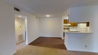 This single-floor unit features an open-concept layout that seamlessly connects the living area, dining space, and kitchen. The living and dining area is finished with durable hardwood-style flooring, while plush carpeting in the adjoining space adds warmth and comfort underfoot. Crisp white walls throughout create a clean, neutral backdrop that lends itself well to a variety of décor styles.

The kitchen is positioned to the side and defined by a white peninsula counter that serves as both a workspace and a natural divider between the cooking and living areas. Warm-toned wood cabinetry lines the upper portion of the kitchen walls, providing ample storage, while a white electric range and ventilation hood complete the functional cooking setup. A dishwasher is also visible, adding convenience to the space.

A doorway visible on the left side of the main living area leads to additional rooms, suggesting a thoughtful floor plan that keeps private spaces separate from the main living zone. The unit is well-lit with ceiling lighting and maintains a compact yet practical layout suited for comfortable everyday living.