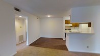 This single-floor unit features an open-concept layout that seamlessly connects the living area, dining space, and kitchen. The living and dining area is finished with durable hardwood-style flooring, while plush carpeting in the adjoining space adds warmth and comfort underfoot. Crisp white walls throughout create a clean, neutral backdrop that lends itself well to a variety of décor styles.

The kitchen is positioned to the side and defined by a white peninsula counter that serves as both a workspace and a natural divider between the cooking and living areas. Warm-toned wood cabinetry lines the upper portion of the kitchen walls, providing ample storage, while a white electric range and ventilation hood complete the functional cooking setup. A dishwasher is also visible, adding convenience to the space.

A doorway visible on the left side of the main living area leads to additional rooms, suggesting a thoughtful floor plan that keeps private spaces separate from the main living zone. The unit is well-lit with ceiling lighting and maintains a compact yet practical layout suited for comfortable everyday living.