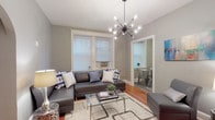 This single-floor residence offers a thoughtfully arranged layout with a cohesive gray color palette and rich hardwood flooring throughout. The living room serves as the centerpiece of the space, anchored by a striking Sputnik-style chandelier that adds a modern touch. Natural light filters in through multiple windows fitted with blinds, illuminating the open feel of the room. A charming arched doorway leads further into the unit, adding a classic architectural detail. The living area flows seamlessly into a separate dining space, visible through a connecting doorway and complete with its own natural light source. The kitchen is equipped with warm-toned wood cabinetry, ample upper and lower storage, a gas range, and generous countertop workspace — all designed with everyday functionality in mind. The unit includes two bedrooms, each featuring the same warm hardwood floors. The primary bedroom benefits from dual windows with a ceiling fan, a classic radiator, and a serene, restful atmosphere. The second bedroom is a quieter, more intimate space accented by contemporary wall art and a bedside lamp. The full bathroom offers a clean, tiled finish with a shower-tub combination and a window for ventilation. Overall, this space combines classic architectural character with modern lighting and a neutral, versatile aesthetic.