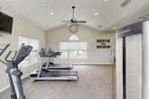 Located at 10010 Skinner Lake Drive in Jacksonville, FL, this fitness center offers a comprehensive workout environment designed to meet a variety of exercise needs. The space is defined by its vaulted ceilings, which lend an open, airy feel throughout, while recessed lighting and ceiling fans ensure comfort during any workout session. Large windows along multiple walls flood the interior with natural light and provide views of the surrounding outdoor environment, creating an inviting and energizing atmosphere. The flooring consists of durable, textured carpet tiles suited for fitness use. The facility is divided into distinct functional zones: a cardio area equipped with Life Fitness treadmills and an elliptical machine, a strength and free weights section featuring a multi-tiered dumbbell rack and adjustable weight bench, and a separate area with cable and resistance machines. A wall-mounted television is positioned for easy viewing during workouts. Full-length mirrors along one wall visually expand the space and support form-checking during exercise. A teal accent wall adds a pop of color, while motivational wall decals reinforce an active mindset. An arched transom window above the entry doors serves as a standout architectural detail, enhancing the overall character of the space.
