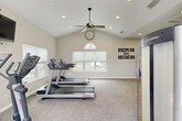 Located at 10010 Skinner Lake Drive in Jacksonville, FL, this fitness center offers a comprehensive workout environment designed to meet a variety of exercise needs. The space is defined by its vaulted ceilings, which lend an open, airy feel throughout, while recessed lighting and ceiling fans ensure comfort during any workout session. Large windows along multiple walls flood the interior with natural light and provide views of the surrounding outdoor environment, creating an inviting and energizing atmosphere. The flooring consists of durable, textured carpet tiles suited for fitness use. The facility is divided into distinct functional zones: a cardio area equipped with Life Fitness treadmills and an elliptical machine, a strength and free weights section featuring a multi-tiered dumbbell rack and adjustable weight bench, and a separate area with cable and resistance machines. A wall-mounted television is positioned for easy viewing during workouts. Full-length mirrors along one wall visually expand the space and support form-checking during exercise. A teal accent wall adds a pop of color, while motivational wall decals reinforce an active mindset. An arched transom window above the entry doors serves as a standout architectural detail, enhancing the overall character of the space.