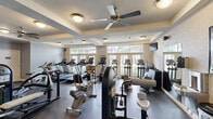 This fitness center in Lafayette, Louisiana, offers a comprehensively outfitted exercise facility across a single floor. The space is thoughtfully designed with tray ceilings accented by ceiling fans, flush-mount lighting, and large multi-pane windows along one wall that invite natural light and outdoor views, creating an open and energizing atmosphere. The durable rubber flooring provides a practical, cushioned surface suitable for a wide range of activities.

The cardio zone is well-stocked with multiple treadmills, elliptical trainers, upright and recumbent stationary bikes, and stair-climbers, all positioned to take advantage of the natural light from the windows. A dedicated strength training area includes a functional trainer cable machine, an Instinct multi-station unit, leg press machines, and various selectorized strength machines. A mirrored wall section expands the sense of space while supporting form checks during workouts. A dumbbell rack with a full range of free weights rounds out the strength offerings. The neutral gray and beige color palette, combined with ample lighting and good air circulation from the ceiling fans, contributes to a clean, focused environment conducive to effective training.