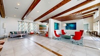 Located at 7995 Haven Way in Kissimmee, Florida, this well-appointed single-floor community clubhouse offers a generous and thoughtfully designed gathering space. The interior showcases a striking mid-century modern aesthetic, anchored by prominent exposed wood beam ceilings that run throughout, large-format tile flooring with wood accent inlays, and crisp white walls that amplify the sense of openness and light.

The expansive living area serves as the heart of the space, featuring a large sectional sofa, a pool table, and a bar counter with pendant lighting and a textured stone backsplash — ideal for socializing and entertainment. A dramatic slatted wood partition wall adds architectural depth, while multiple flat-screen televisions are positioned throughout for convenience.

The lounge area is furnished with bold red accent chairs arranged around a low coffee table, accented by vibrant large-scale artwork and a decorative wood ceiling trellis. A separate, more relaxed seating zone features a teal sectional sofa, geometric wall art, and a large mounted Scrabble board, adding a playful, community-oriented touch.

The dining room provides a dedicated meeting space equipped with a glass-top conference table, ergonomic mesh chairs, a wall-mounted display screen, and a striking gold starburst chandelier overhead. Natural light filters in through large windows, offering views of the surrounding greenery and enhancing the room's welcoming atmosphere.