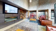 Located on Commerce Drive in Exton, PA, this single-floor amenity space showcases a striking industrial aesthetic that blends raw materials with thoughtfully curated design. Soaring ceilings clad in warm wood planking contrast with exposed brick walls, textured concrete surfaces, and polished terrazzo floors that extend throughout the space.

The grand open lobby greets visitors with floor-to-ceiling steel-framed windows that flood the interior with natural light and frame views of an outdoor patio furnished with seating and a fire pit. Leather sofas and lounge chairs are arranged across layered area rugs, anchored by bold vintage-industrial décor, including large red industrial gear wheels used as sculptural accents.

A dedicated lounge area features a large projection screen, built-in shelving, and additional leather seating. Nearby, curved leather booth seating with reclaimed wood detailing sits beneath an artistic pipe-and-gauge wall installation. A game area includes a shuffleboard table, while a kitchenette offers reclaimed wood cabinetry, subway tile, and a coffee station alongside a large antique press sculpture.

A conference area provides a long communal table with rolling leather chairs beneath pendant lighting. An elevator alcove with decorative metal mesh screening completes the space.