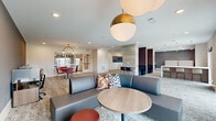 This thoughtfully designed community lounge and co-working space spans a single, open floor plan, offering a variety of distinct functional zones within a cohesive, modern aesthetic. The space is finished with patterned, neutral-toned carpet throughout, contributing to a warm yet polished atmosphere.

Upon entering through glass-paneled doors, the layout opens into a spacious lounge area anchored by a curved, dark upholstered sofa and coordinating seating, accented by striking gold globe pendant lights overhead. A wood-paneled accent column adds an organic warmth that contrasts with the crisp white walls and ceilings.

A counter-height bar runs along one wall, outfitted with slim stools in rich burgundy tones, ideal for casual individual work or conversation. Nearby, a central high-top table with padded stools sits beneath a cluster of brushed gold pendant lights, bathed in natural light from large windows overlooking an outdoor streetscape.

A dedicated dining and gathering nook features round wooden tables surrounded by dusty rose upholstered chairs, illuminated by an elegant arched gold pendant fixture. This area is accented by a bold triptych of vintage typewriter artwork, lending a creative, editorial character to the space.

A workstation area with desktop computers rounds out the functionality of this versatile community hub, making it equally suited for focused work, socializing, or collaborative gatherings.