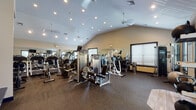 This single-floor fitness center is thoughtfully designed to support a full range of workout routines. The space is defined by its distinctive vaulted ceilings, which lend an open, airy quality to the room and are complemented by recessed lighting and ceiling fans that maintain a comfortable environment during workouts. Durable, dark rubber flooring covers the main exercise area, providing both safety and practicality underfoot.

The main floor is generously outfitted with a variety of strength-training machines, including cable and weight stack systems suited for both upper and lower body exercises. A dedicated section houses cardio equipment, including treadmills, elliptical trainers, and stair climbers, positioned near windows that allow natural light to filter in and offer views of the surrounding greenery. Full-length wall mirrors expand the visual space and allow users to monitor their form throughout their session.

A functional training zone features a medicine ball rack with multiple weighted options, a stability ball, and a dedicated open floor area for stretching or bodyweight exercises. Wall-mounted televisions provide entertainment, while a row of coat hooks and a bench offer convenient space to prepare before and after workouts. Large windows with blinds frame outdoor views, connecting the interior to the natural surroundings. The overall ambiance is clean, practical, and inviting for regular fitness use.
